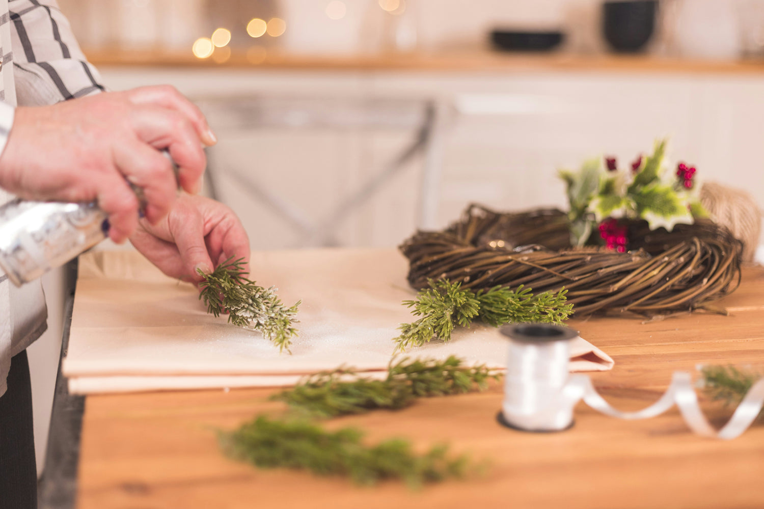 A hand spraying white snow effect onto sprigs of fir, making a handmade Christmas wreath